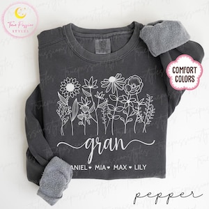 May include: A dark gray sweatshirt with a white floral design and the word "gran" in script. Below the word "gran" are the names Daniel, Mia, Max, and Lily. The sweatshirt has a fleece lining and the text "Comfort Colors" is visible.