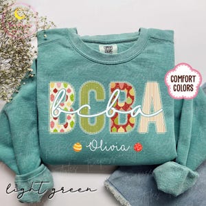 May include: Light green sweatshirt with the letters "BCBA" in a decorative font. The letters are filled with patterns of strawberries, green, and red ornaments. The name "Olivia" is written below the letters.