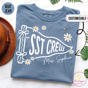 Può includere: Una t-shirt color blu jeans con una grafica bianca a forma di gagliardetto che riporta il testo SST CREW e Mrs. Sophia. Il design include margherite bianche con centri gialli e un palo decorativo con fiocchi sul lato sinistro del gagliardetto.
