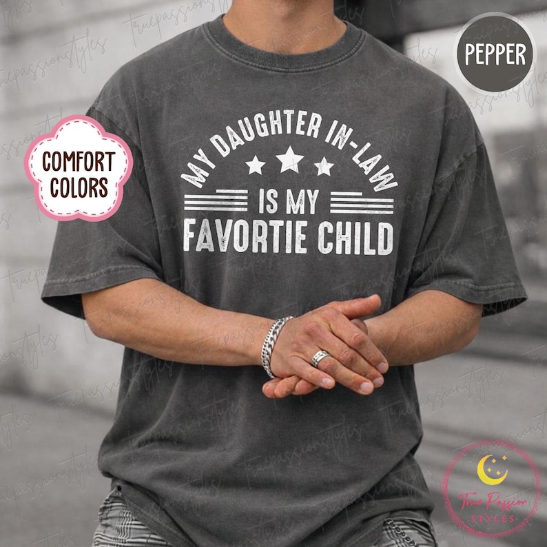 May include: A dark grey t-shirt with the white text "MY DAUGHTER IN-LAW IS MY FAVORITE CHILD". The shirt has a crew neck and short sleeves. The shirt is a comfort color.