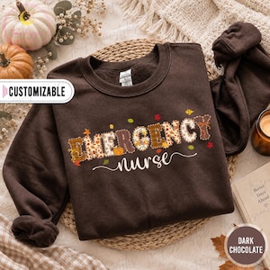 May include: A dark chocolate brown crewneck sweatshirt featuring the text "EMERGENCY" in large, patterned block letters with autumn-themed motifs like pumpkins and leaves, followed by the word "nurse" in a white cursive script font.