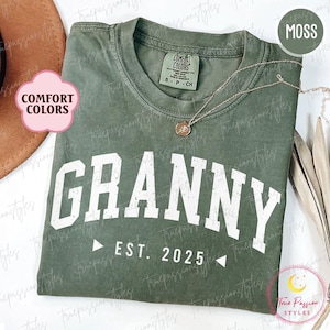 May include: A moss green t-shirt with the text "GRANNY EST. 2025" in white distressed lettering. The shirt is made of a soft, comfortable fabric.