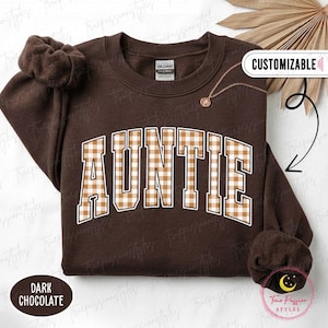 May include: A dark chocolate brown crewneck sweatshirt featuring the word AUNTIE in a white-outlined, tan and white gingham plaid collegiate font. The garment includes a matching brown scrunchie and a gold-tone necklace with a small pendant.