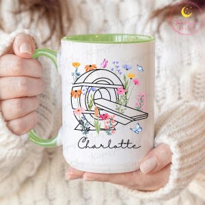 May include: White ceramic mug with a green handle and interior. The mug features a black line drawing of a CT scanner surrounded by colorful flowers and butterflies. The name "Charlotte" is written in cursive below the scanner.