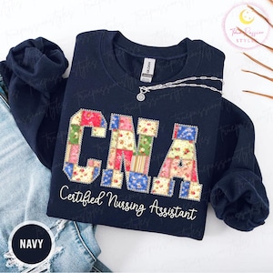 Peut inclure: Sweat-shirt bleu marine avec les lettres "CNA" dans un motif patchwork. Les mots "Certified Nursing Assistant" sont imprimés en dessous en blanc. Un collier argenté est drapé sur le sweat-shirt.