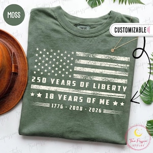 May include: A moss green t-shirt featuring a distressed white graphic of an American flag. The text reads 250 YEARS OF LIBERTY, 18 YEARS OF ME, and the years 1776 - 2008 - 2026. The design includes stars and horizontal stripes in a weathered style.