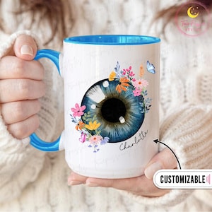 May include: White ceramic mug with a blue interior and handle, featuring a detailed blue eye design surrounded by colorful flowers and a butterfly. The name "Charlotte" is written on the mug. The mug is customizable.