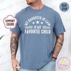May include: A blue jean-coloured t-shirt with the white text "MY DAUGHTER IN-LAW IS MY FAVORITE CHILD". The shirt has a crew neck and short sleeves. The words "COMFORT COLORS" and "BLUE JEAN" are also visible.