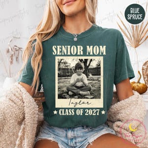 Może przedstawiać: Tealowa koszulka z białym napisem "SENIOR MOM". Poniżej znajduje się czarno-białe zdjęcie dziecka. Zdjęcie jest podpisane "Taylor" i "CLASS OF 2027". Koszulka ma luźny kr&oacute;j.