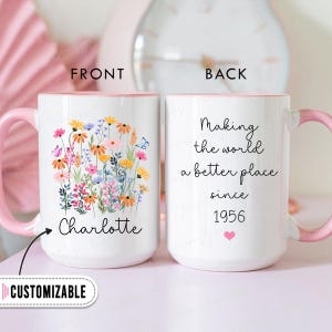 May include: White ceramic mug with a pink handle and interior. The front features a floral design with the name "Charlotte". The back of the mug reads "Making the world a better place since 1956" with a pink heart.