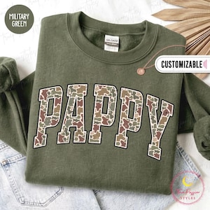 May include: A military green crewneck sweatshirt featuring the word PAPPY in large block letters with a camouflage pattern. The text has a black outline. A small circular tag with the text CUSTOMIZABLE is visible near the collar area.