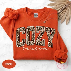 May include: An orange crewneck sweatshirt featuring the word COZY in large block letters with a brown and black leopard print pattern, and the word season written below in white cursive script. The garment has long sleeves with ribbed cuffs.
