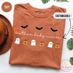 May include: A rust-coloured crew neck t-shirt featuring a graphic print of three black bats, three white ghosts, and three orange jack-o'-lanterns. The text "mother baby nurse" is printed in white cursive script across the centre of the shirt.
