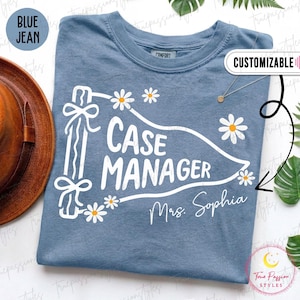 May include: A blue jean colored t-shirt featuring a white graphic of a pennant flag with the text "CASE MANAGER" and "Mrs. Sophia" written below. The design includes white daisy flowers with yellow centers and a ribbon tied to the flag pole.
