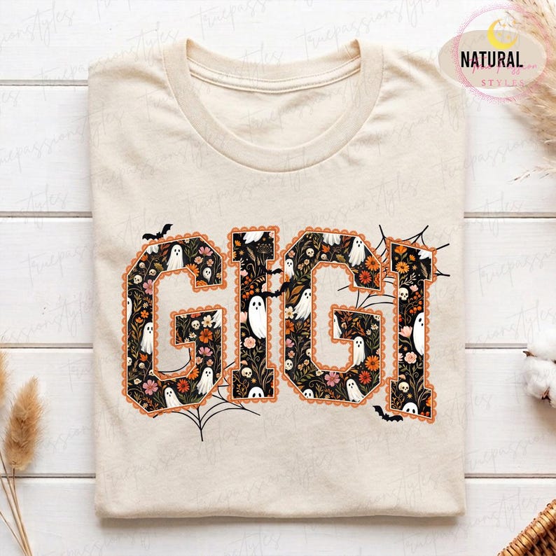 K&ouml;nnte beinhalten: Ein cremefarbenes T-Shirt mit dem Schriftzug GIGI in gro&szlig;en Blockbuchstaben. Die Buchstaben sind mit einem Muster aus wei&szlig;en Geistern, kleinen Totenk&ouml;pfen sowie orangefarbenen und rosafarbenen Blumen auf schwarzem Grund gef&uuml;llt, erg&auml;nzt durch Spinnenweben und Flederm&auml;use.