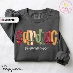May include: A dark gray crewneck sweatshirt featuring the text "CARDIAC" in large, multicolored letters intertwined with a string of Christmas lights, and the word "sonographer" in white script below. A "CUSTOMIZABLE" label is visible.