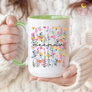 Könnte beinhalten: Weißer Keramikbecher mit grünem Griff und Innenraum. Der Becher ist mit einem farbenfrohen Blumendesign und dem Wort "meemaw" in schwarzer Schrift verziert. Die Blumen umfassen rosa, gelbe und blaue Blüten.