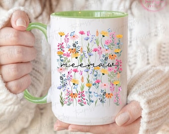 May include: White ceramic mug with a green handle and interior. The mug is decorated with a colorful floral design and the word "meemaw" in black script. The flowers include pink, yellow, and blue blooms.