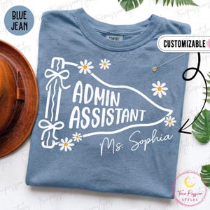 Op de afbeelding: Een blauw spijkerkleurig t-shirt met een witte afbeelding van een wimpel met de tekst "ADMIN ASSISTANT" en "Ms. Sophia" eronder. Het ontwerp bevat witte madeliefjes met gele harten en een lint dat aan de vlaggenmast is gebonden.