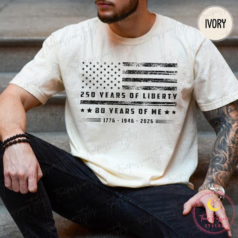 May include: Cream-coloured t-shirt with a distressed American flag graphic. Text includes "250 YEARS OF LIBERTY" and "80 YEARS OF ME", with the years 1776-1946-2026. The shirt is a casual style.