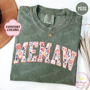 May include: A moss green Comfort Colors t-shirt with the word "MEMAW" in a floral design. The shirt has a crew neck and short sleeves. A gold necklace with a pendant is draped on the shirt.
