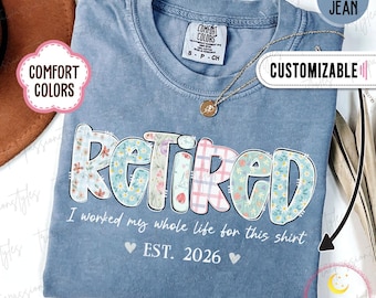 May include: Blue jean-colored t-shirt with the word "RETIRED" in a floral design. The shirt also has the text "I worked my whole life for this shirt" and "EST. 2026". A gold necklace is visible.