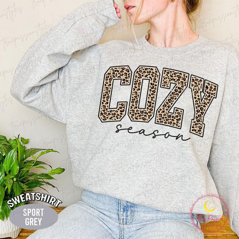 May include: A light gray crewneck sweatshirt featuring the word COZY in large block letters with a brown and black leopard print pattern. Below the word COZY, the word season is printed in a black cursive script font. The sweatshirt has long sleeves.