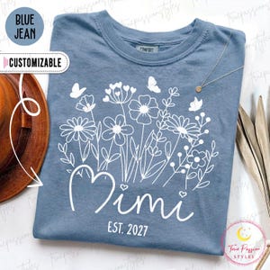 Könnte beinhalten: Ein blaues T-Shirt mit einem weißen Grafikdruck aus verschiedenen Wildblumen und Schmetterlingen. Der Text "Mimi" ist in Schreibschrift über dem Text "EST. 2027" in einer serifenlosen Schriftart gedruckt. Das Shirt hat einen Rundhalsausschnitt.
