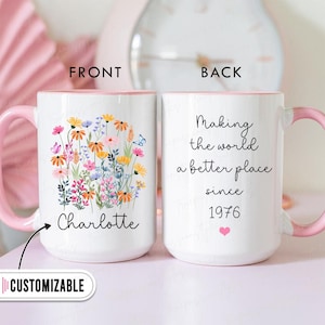 Peut inclure: Mug en céramique blanche avec une anse et un intérieur roses. L'avant présente un motif floral avec le nom "Charlotte". Le dos indique "Making the world a better place since 1976" avec un cœur rose. La tasse est personnalisable.