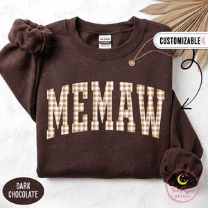 Könnte beinhalten: Ein dunkelbraunes Sweatshirt mit Rundhalsausschnitt und dem Wort MEMAW in einem beigefarbenen und weißen Vichy-Karomuster auf der Brust. Das Kleidungsstück hat lange Ärmel mit gerafften Bündchen und ein Gildan-Markenetikett am inneren Halsausschnitt.