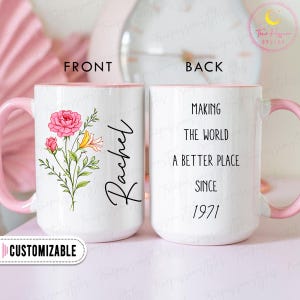 Puede incluir: Taza de cerámica blanca con interior y asa rosas. La parte delantera presenta un diseño floral con rosas rosas y lirios amarillos, el nombre "Rachel" y la palabra "CUSTOMIZABLE". La parte trasera dice "MAKING THE WORLD A BETTER PLACE SINCE 1971".
