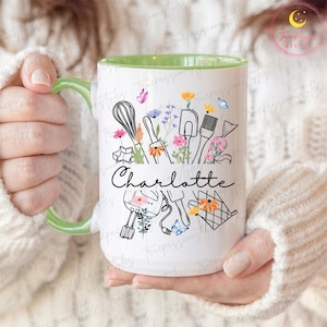 May include: White ceramic mug with a green handle and interior. The mug features a black line drawing of baking tools and flowers, with the name "Charlotte" in script. Butterflies and floral accents add color.