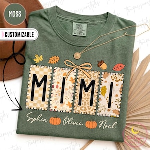 Könnte beinhalten: Ein moosgrünes T-Shirt mit dem Wort MIMI in großen schwarzen Buchstaben, wobei jeder Buchstabe vor einem unterschiedlich gemusterten rechteckigen Hintergrund steht. Das Design enthält eine beige Schleife, Herbstblätter, Eicheln und die Namen Sophia, Olivia und Noah.