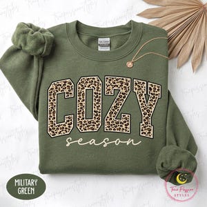 May include: A military green crewneck sweatshirt featuring the word COZY in large block letters with a brown and black leopard print pattern, and the word season written below in a white cursive script. A gold chain necklace rests on the fabric.