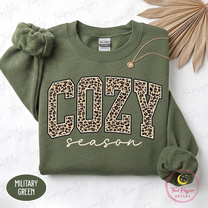 May include: A military green crewneck sweatshirt featuring the word COZY in large block letters with a brown and black leopard print pattern, and the word season written below in a white cursive script. A gold chain necklace rests on the fabric.
