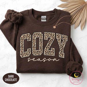 May include: A dark chocolate brown crewneck sweatshirt featuring the word COZY in large block letters with a tan and black leopard print pattern, and the word season written below in a white cursive script. The garment has a Gildan brand label.
