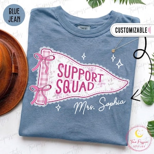 May include: A blue jean colored t-shirt featuring a graphic of a pennant flag with pink gingham trim, two pink bows, and a floral pattern. The text reads SUPPORT SQUAD and Mrs. Sophia in white and pink script. The design includes small white star icons.