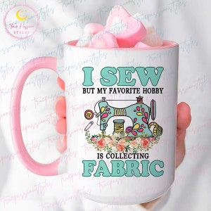 Peut inclure: Une tasse en céramique blanche avec une poignée rose. La tasse présente une illustration colorée d'une machine à coudre avec des fleurs et le texte "Je couds mais mon passe-temps préféré est de collectionner du tissu".