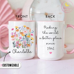 Op de afbeelding: Witte keramische mok met een roze handvat en binnenkant. De voorkant heeft een bloemenontwerp met de naam "Charlotte" en het woord "AANPASBAAR". De achterkant leest "Making the world a better place since 1941".