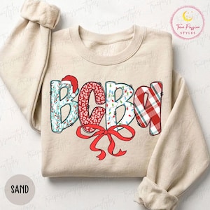 May include: A sand-colored crewneck sweatshirt featuring the letters BCBA in a festive design. The letters are decorated with holiday patterns including holly leaves, red leopard print, Christmas lights, and a red plaid pattern with a red bow.