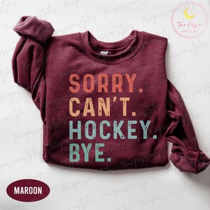 May include: A maroon crew neck sweatshirt with the text "SORRY. CAN'T. HOCKEY. BYE." in orange, tan, and teal lettering. The sweatshirt has long sleeves and a ribbed neckline.