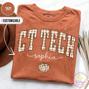 May include: A rust-colored short-sleeve t-shirt featuring the text CT TECH in a brown and white gingham pattern. Below the text, the name sophia is printed in a white cursive font, followed by a small pumpkin graphic with a gingham pattern.