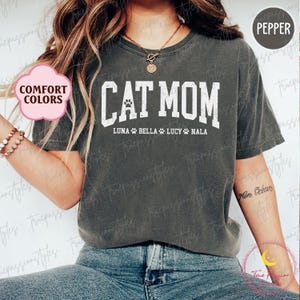 May include: A pepper-coloured t-shirt with the white text "CAT MOM" and a paw print graphic. Underneath are the names "LUNA BELLA LUCY NALA". The shirt is from the "Comfort Colors" range.