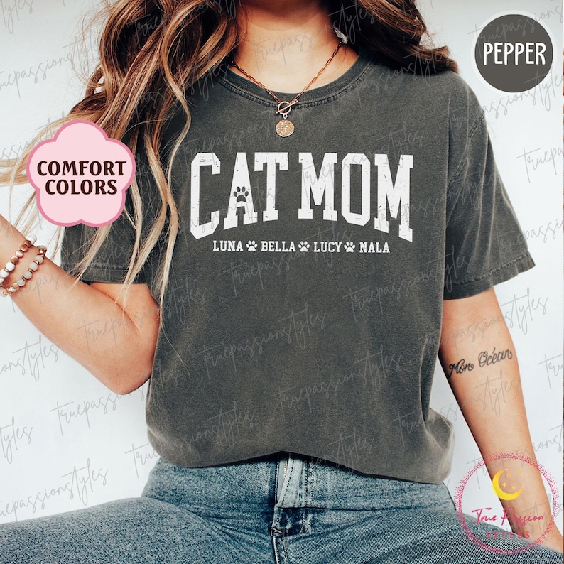 May include: A pepper-coloured t-shirt with the white text "CAT MOM" and a paw print graphic. Underneath are the names "LUNA BELLA LUCY NALA". The shirt is from the "Comfort Colors" range.