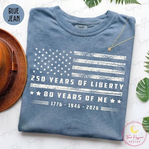 May include: Blue jean-coloured t-shirt with a distressed American flag design. The shirt features the text "250 YEARS OF LIBERTY" and "80 YEARS OF ME" with the years 1776-1946-2026. The shirt has a crew neck and a comfort fit.