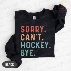 May include: Black crew neck sweatshirt with the text "SORRY. CAN'T. HOCKEY. BYE." in a retro font. The text is in red, orange, teal, and white. The word "BLACK" is at the bottom of the sweatshirt.
