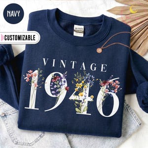 May include: A navy blue crewneck sweatshirt featuring the white text VINTAGE 1946. The numbers are decorated with colorful floral illustrations including pink, purple, yellow, and blue wildflowers. The garment has a Gildan brand label at the neck.