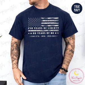 May include: Navy blue t-shirt featuring a distressed American flag design. The graphic includes the text "250 YEARS OF LIBERTY" and "80 YEARS OF ME" with the years 1776-1946-2026. The shirt has a crew neck.