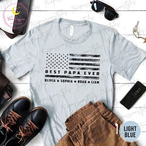 K&ouml;nnte beinhalten: Hellblaues T-Shirt mit einem verblassten amerikanischen Flaggen-Design. Das Shirt tr&auml;gt den Text "BEST PAPA EVER" und die Namen Olivia, Sophia, Noah und Liam. Das Shirt wird mit brauner Hose, schwarzen Schuhen und Sonnenbrille gezeigt.