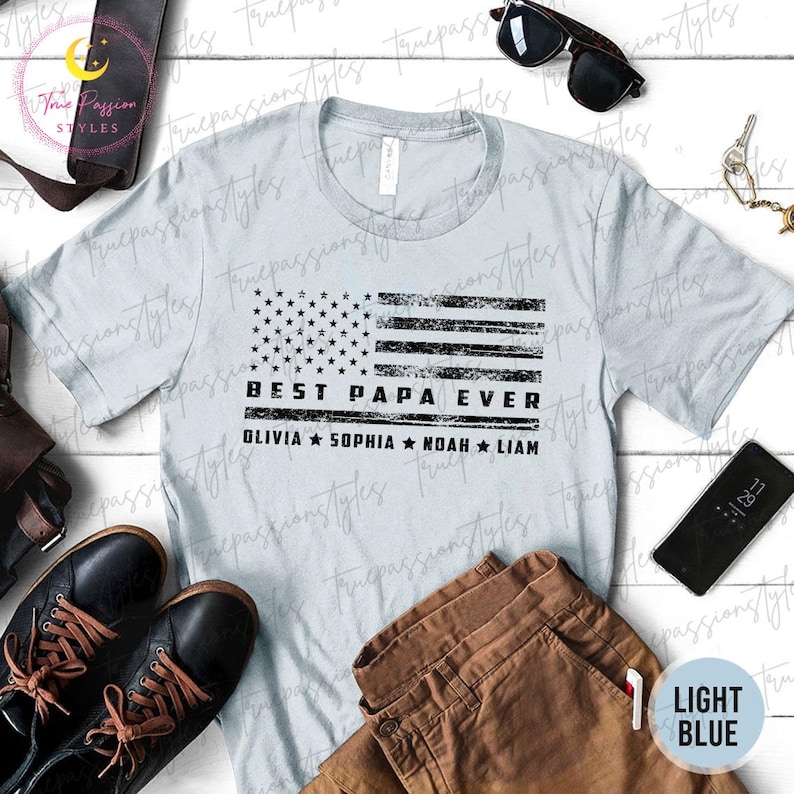 K&ouml;nnte beinhalten: Hellblaues T-Shirt mit einem verblassten amerikanischen Flaggen-Design. Das Shirt tr&auml;gt den Text "BEST PAPA EVER" und die Namen Olivia, Sophia, Noah und Liam. Das Shirt wird mit brauner Hose, schwarzen Schuhen und Sonnenbrille gezeigt.